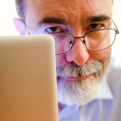 A man with grey beard and glasses, looking at the camera, from behind a laptop screen